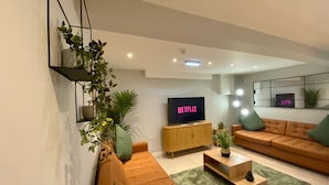 Una sala de estar moderna con un sofá seccional, un televisor mostrando 'NETFLIX' y un estante montado en la pared con plantas.