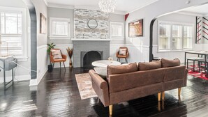 A modern living room with a fireplace, a brown sofa, and a chandelier. - 15 mins to beach/Renew+Relax/Clean/ Nat’l Park!/Theater/Game Room/Pet Friendly (Gary)