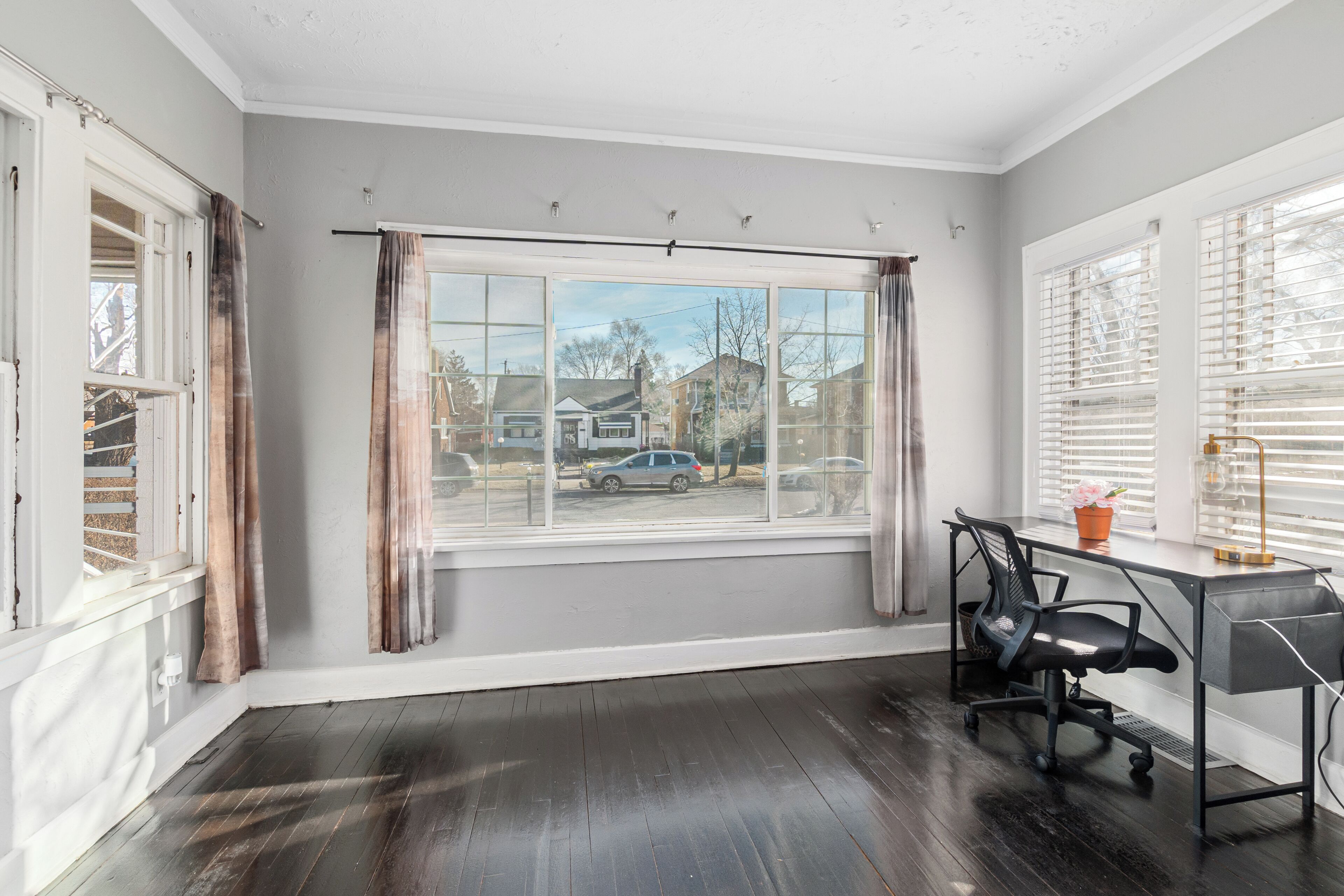A room with a large window, a desk with a chair, and a view of a residential street.
