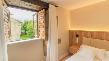 Un dormitorio con cama, cabecera de madera, ventana con contraventanas abiertas y vista a un jardín.