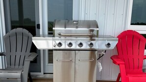 Una parrilla de acero inoxidable para exteriores con cuatro quemadores, ubicada en una terraza de madera con dos sillas Adirondack, una gris y otra roja, frente a una puerta blanca.