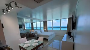 Une chambre d’hôtel moderne dotée d’un grand lit, d’un coin repas avec une table dressée pour deux personnes, et offrant une vue sur la mer grâce à de grandes fenêtres.