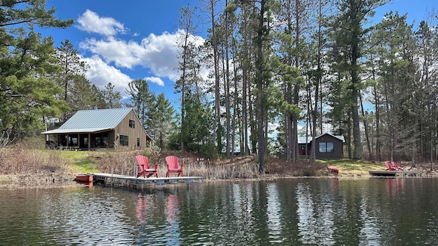 En stuga vid sjön med två röda Adirondack-stolar på en brygga, omgiven av höga träd och en klar himmel.
