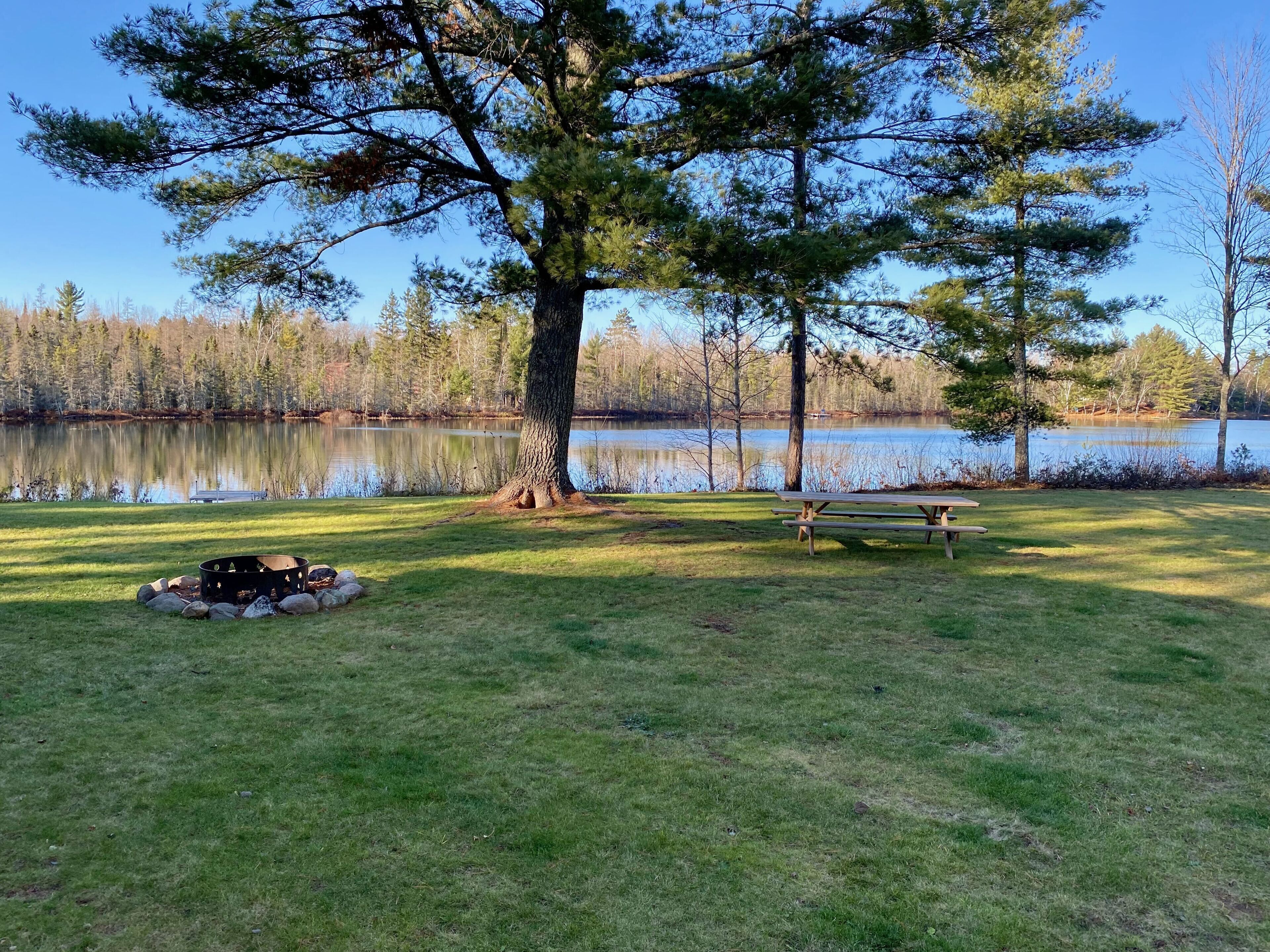 Ein Picknickplatz mit Feuerstelle und Bänken am See, umgeben von Bäumen.