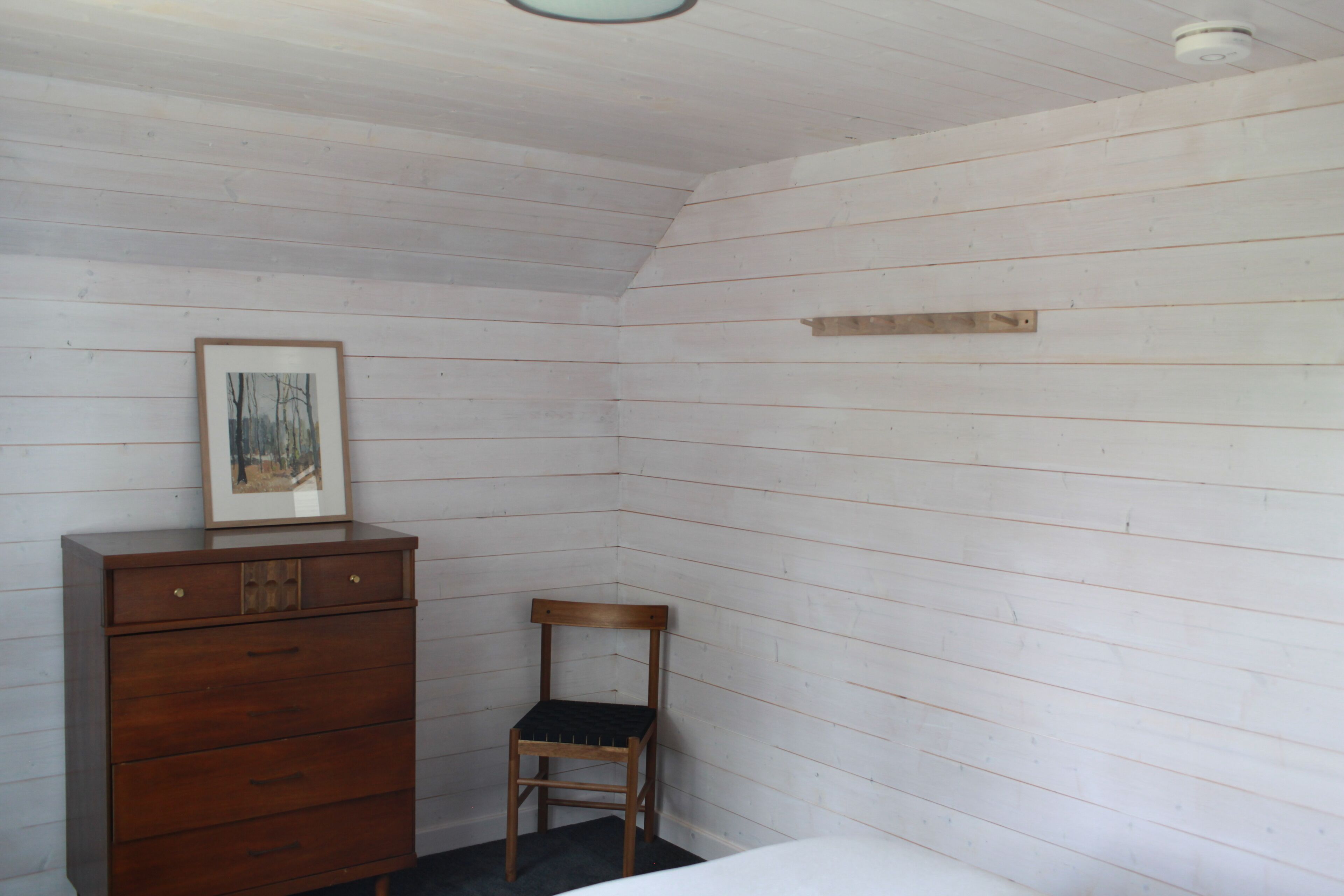 A room with white wooden walls, a wooden dresser with drawers, a framed picture on top, and a wooden chair.