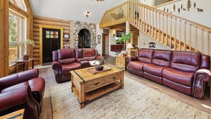 A log cabin interior with a stone fireplace, leather sofas, a wooden coffee table, and a staircase with a wooden railing. - Hawk's Landing | A Refined Modern Mountain Escape (Cascade-Chipita Park)