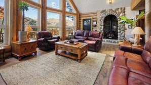 A spacious living room with large windows, a stone fireplace, leather sofas, and a wooden coffee table. - Hawk's Landing | A Refined Modern Mountain Escape (Cascade-Chipita Park)
