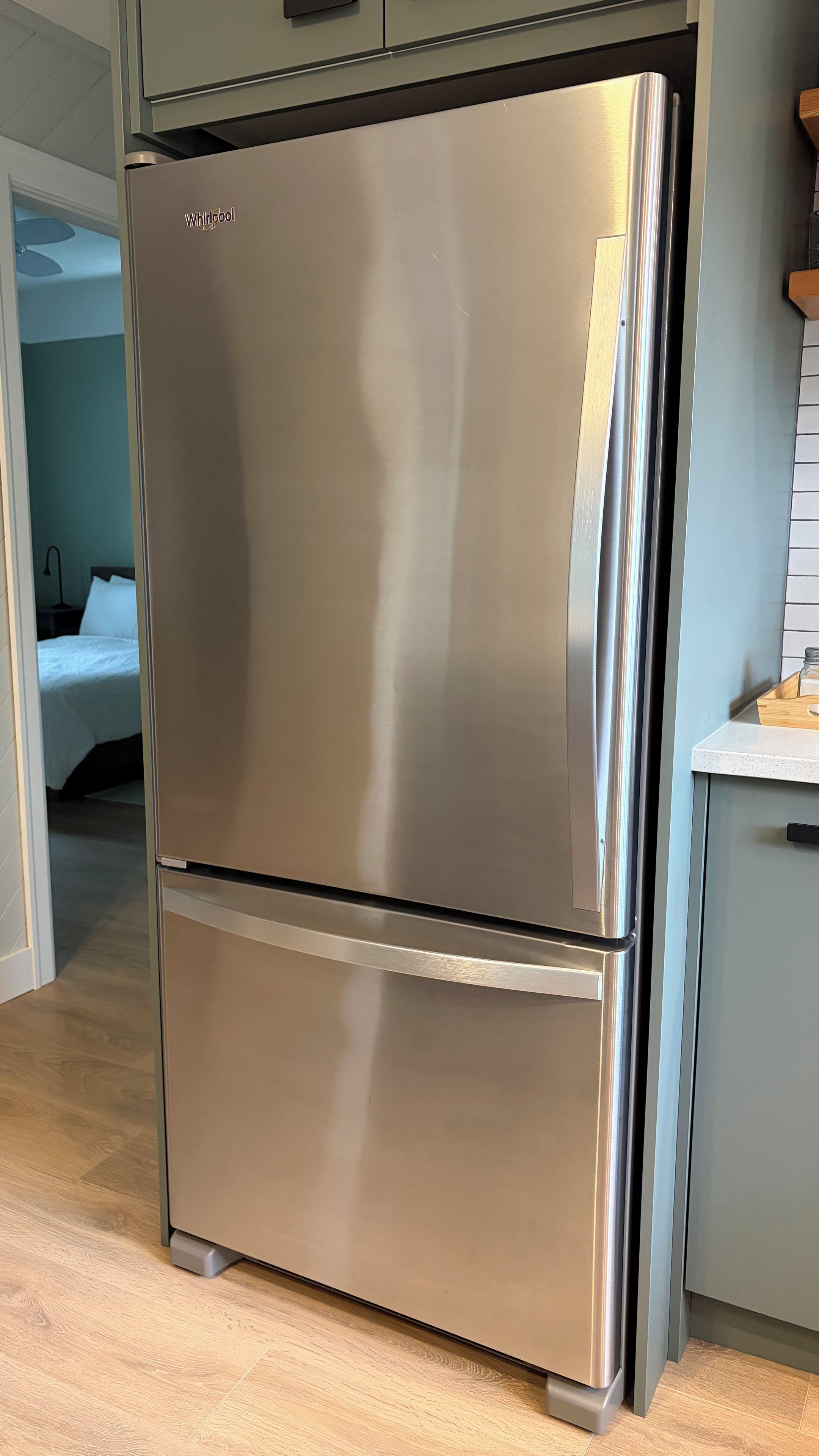 Uma geladeira Whirlpool de aço inoxidável em uma cozinha com piso de madeira e um armário cinza.