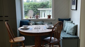 A dining area with a round wooden table, two wooden chairs, a bench with cushions, and a window with potted plants. - Charming Cottage Centrally Located
Business Licence #28949 (Victoria)