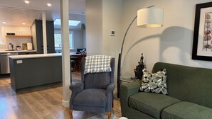A modern living room with a green sofa, a grey armchair, and a floor lamp. A kitchen with a dining area is visible in the background. - Charming Cottage Centrally Located
Business Licence #28949 (Victoria)