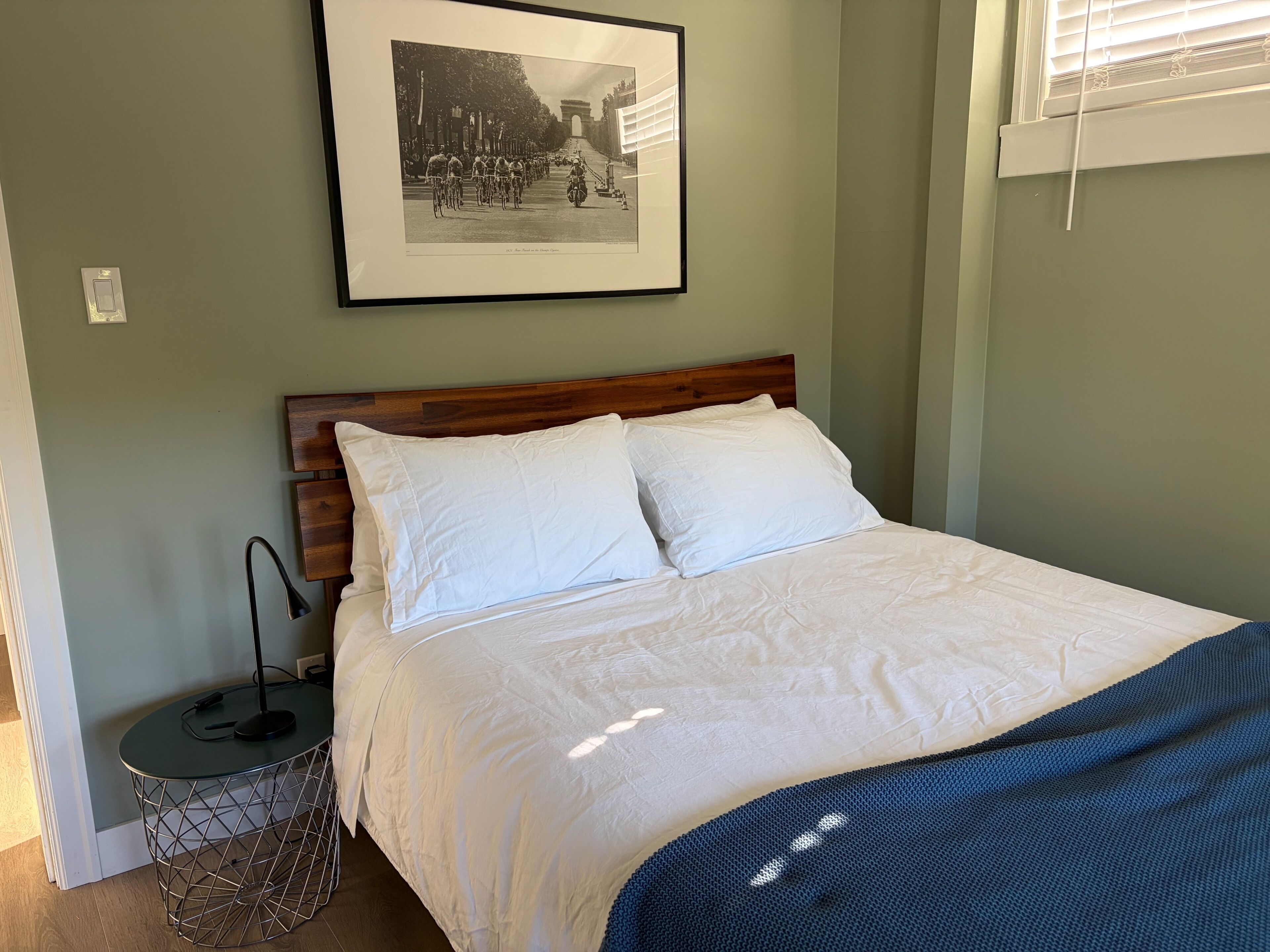 Un dormitorio con cama, almohadas, cabecero, un cuadro en la pared, una mesita de noche con lámpara y una ventana con persianas.