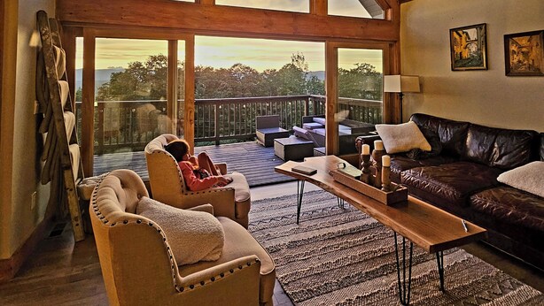 Un salon spacieux avec un haut plafond, de grandes fenêtres, une terrasse en bois et un coin salon confortable.
