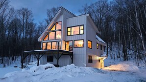 Une maison moderne à deux étages, dotée d’un grand porche, entourée d’un paysage enneigé et d’une forêt dense.