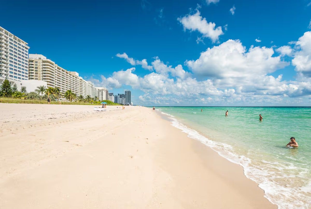 Une plage avec un rivage sablonneux et une eau d’un bleu limpide, avec des bâtiments en arrière-plan.