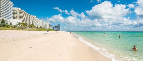 Una playa con orilla arenosa y agua azul cristalina, con edificios al fondo.