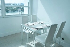 A dining area with a table set for four, white chairs, and a view of a body of water through a window. - BEACHFRONT CONDO-HOTEL bayview with free parking (Miami)