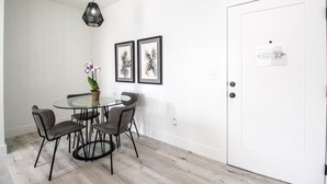 Un comedor con mesa de vidrio y cuatro sillas, una planta en maceta y dos cuadros en la pared.
