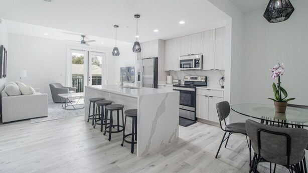 Una cocina moderna con una isla central, taburetes de bar y un comedor con una mesa de vidrio y sillas.