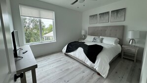 Un dormitorio con cama, mesitas de noche, escritorio, ventilador de techo y ventana con vistas a árboles.