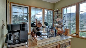 A kitchen with a Keurig coffee maker, various pots and pans, a hanging rack, and a view of a mountainous landscape through large windows. - Raven's Nest | Modern Forest Sanctuary (Cascade)