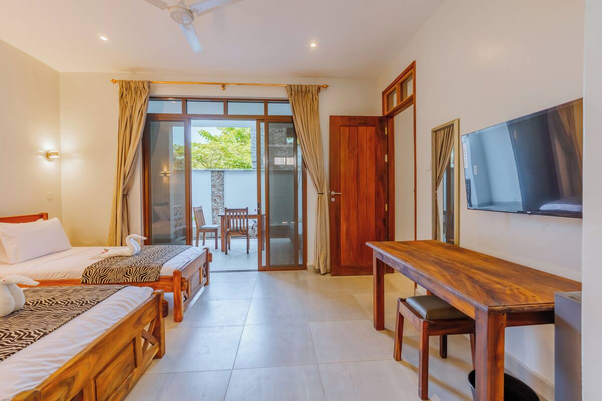 A hotel room with two beds, a wooden table, a TV, and a sliding glass door leading to an outdoor area.
