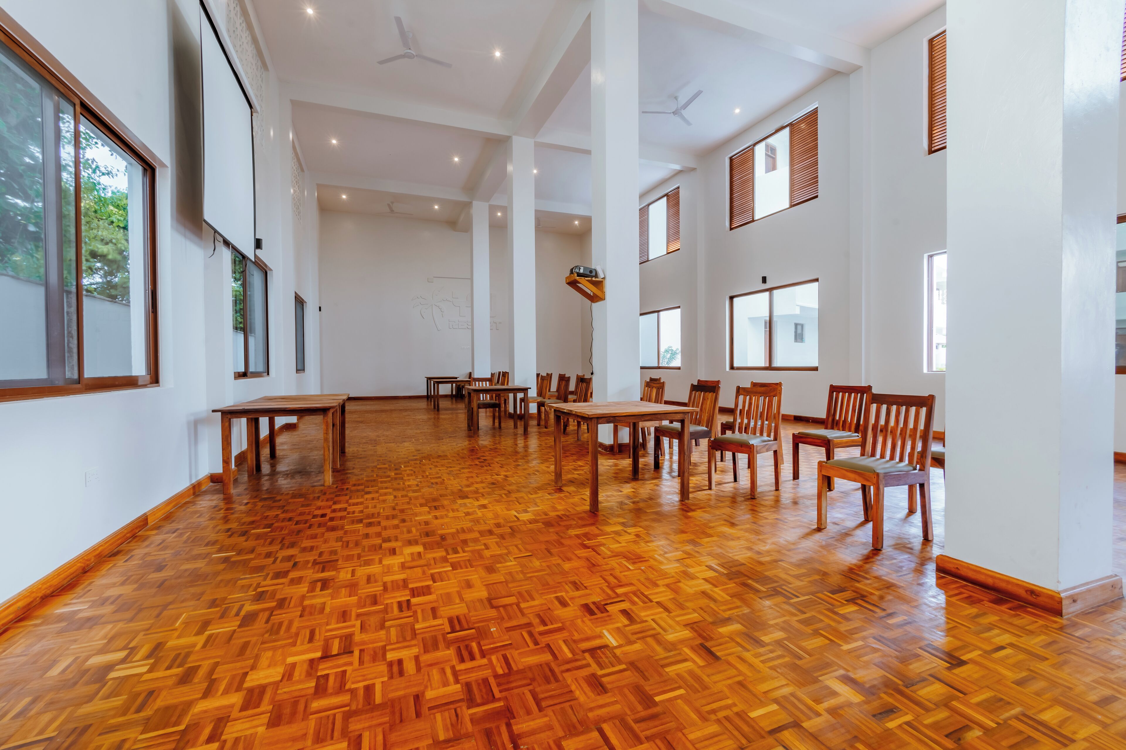 A spacious room with a wooden parquet floor, multiple tables and chairs, large windows, and ceiling fans.