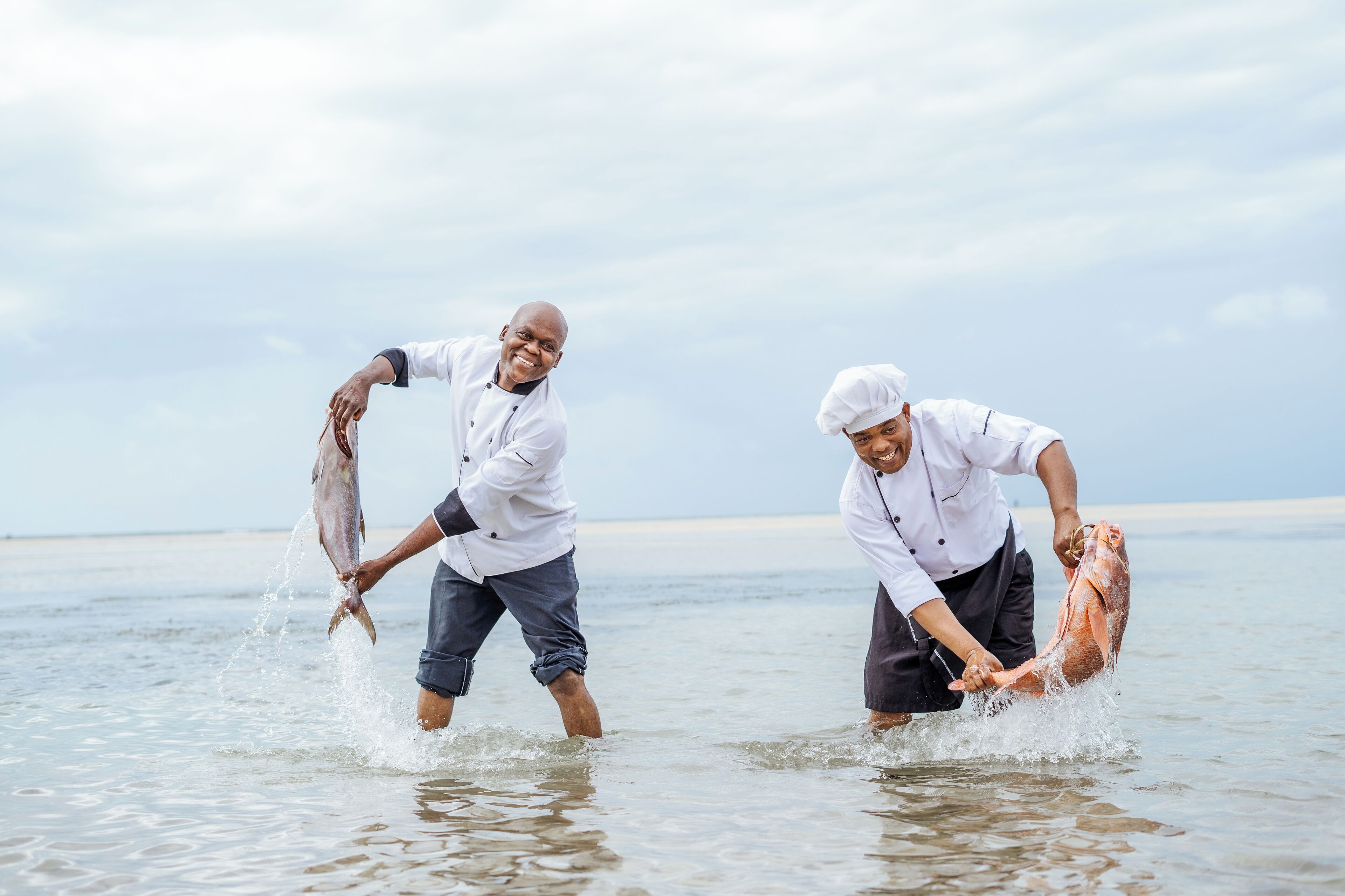Dos personas con uniforme de chef sostienen pescado en agua poco profunda.