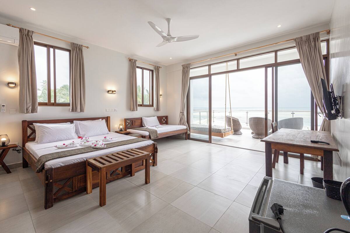 A hotel room with two beds, a dining table, a ceiling fan, and large windows with curtains.