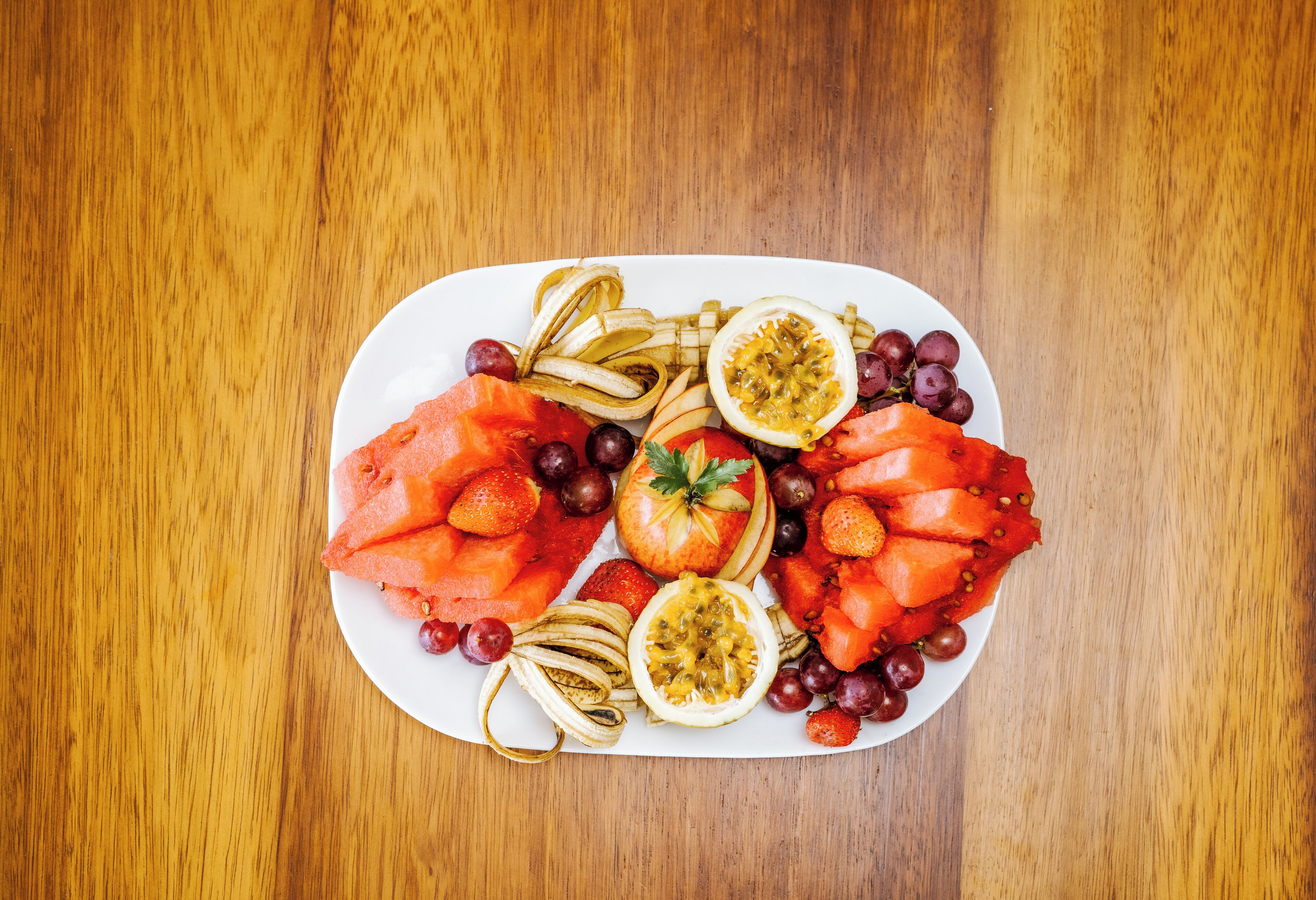 Een bord met verschillende soorten fruit, zoals watermeloen, aardbeien, druiven en passievrucht, op een houten tafel.