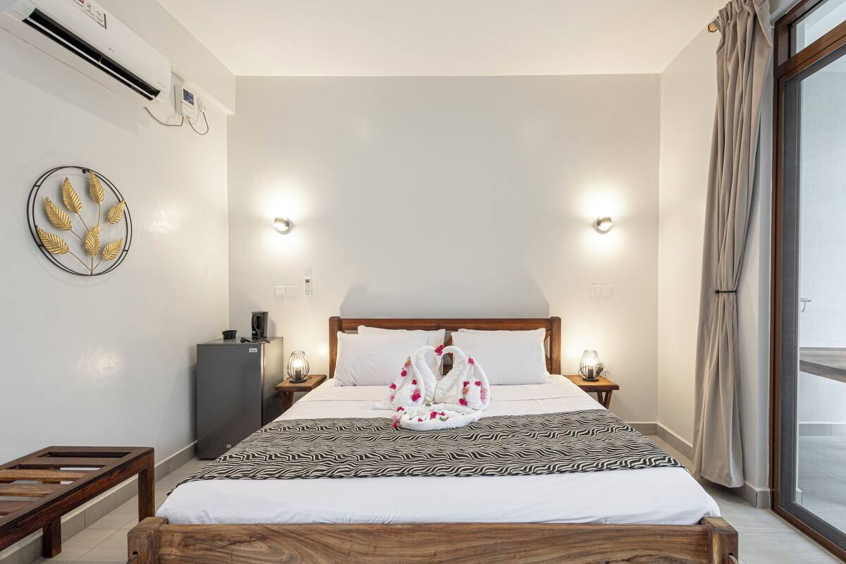 A hotel room with a bed, a small desk, a refrigerator, and a wall-mounted air conditioner.