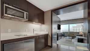 Una cocina moderna con un microondas Bosch, gabinetes de acero inoxidable, un fregadero y un separador de vidrio que conduce a una zona de estar con un sofá y un televisor.
