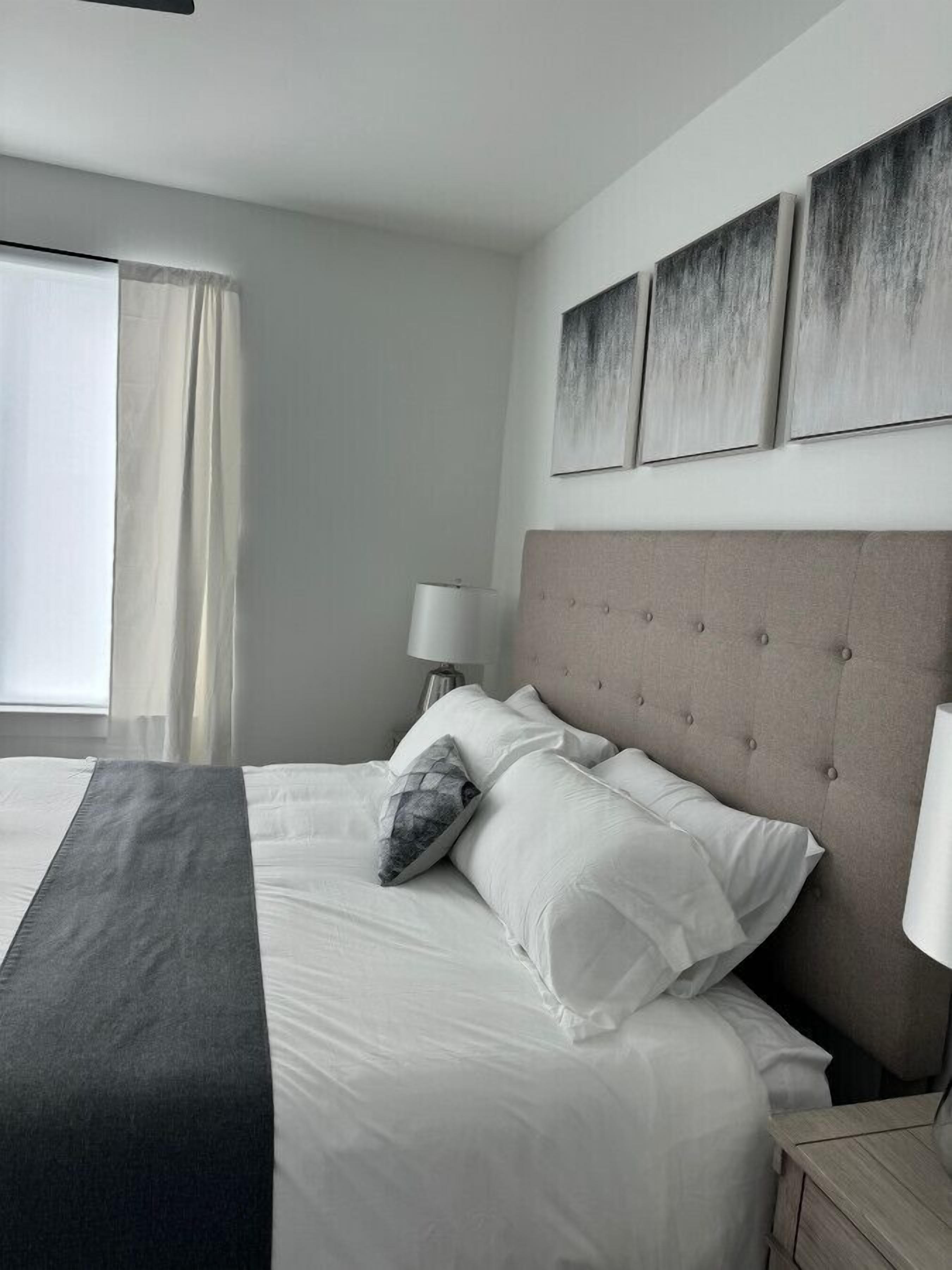 Una habitación de hotel moderna con una cama grande, cabecera con botones, mesita de noche con lámpara y ventana con cortinas.