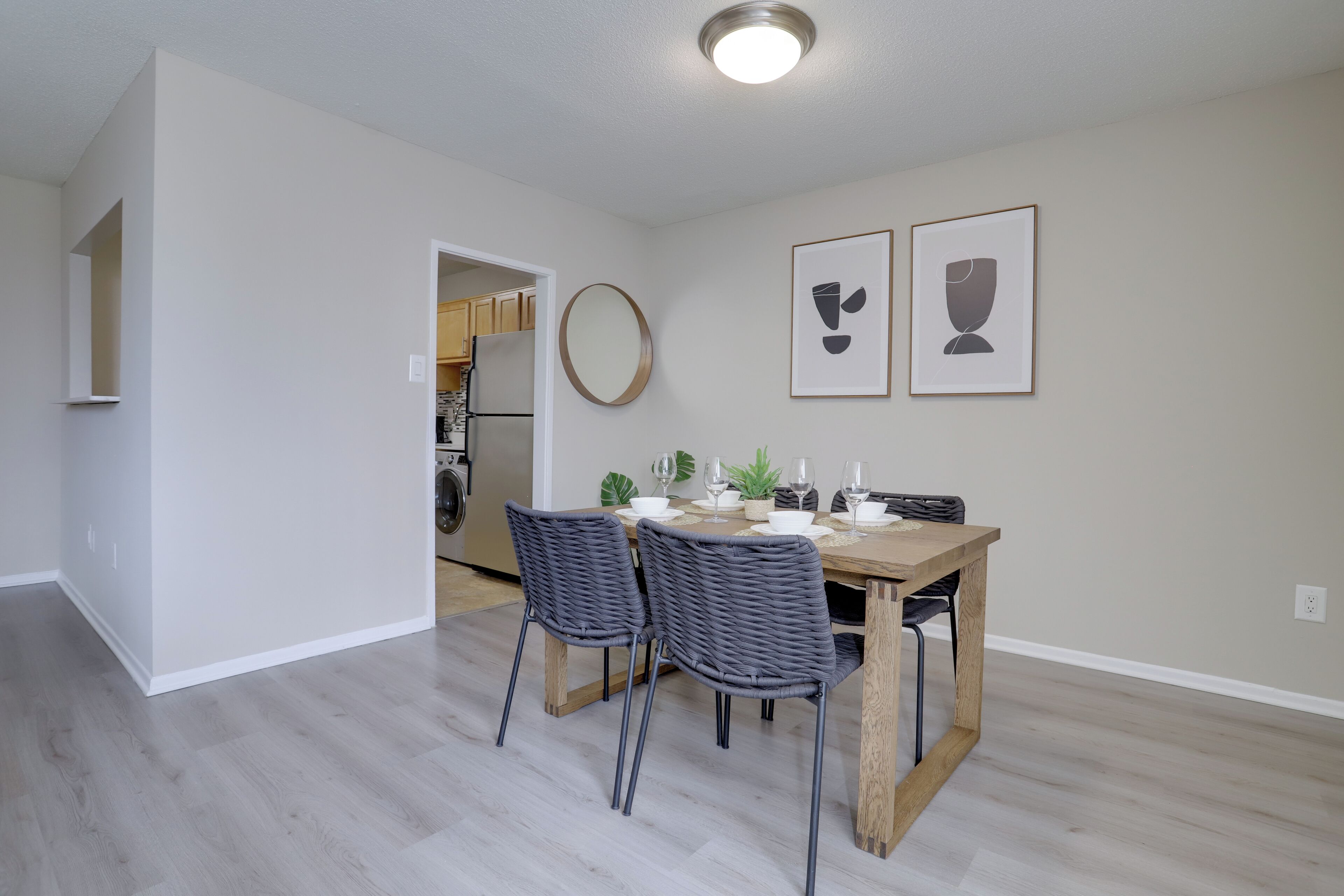 A dining area with a wooden table and two chairs, a kitchen in the background, and two framed pictures on the wall.