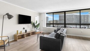 A modern living room with a large window, a flat-screen TV, a black sofa, a wooden console, and a floor lamp. - Comfort Living Alexandria Apts (Arlington)