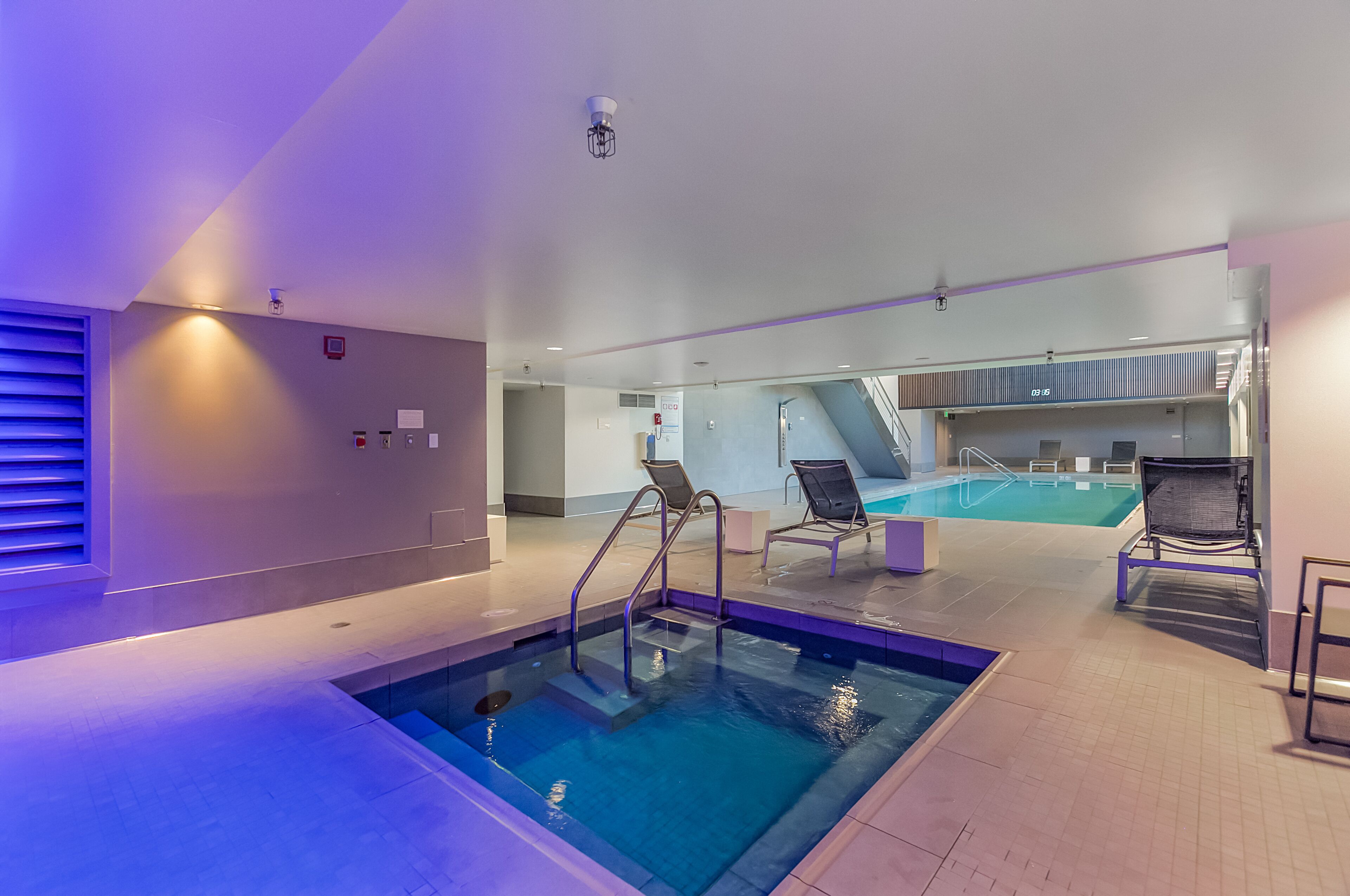 An indoor swimming pool with clear water, surrounded by lounge chairs and a staircase leading to an upper level.