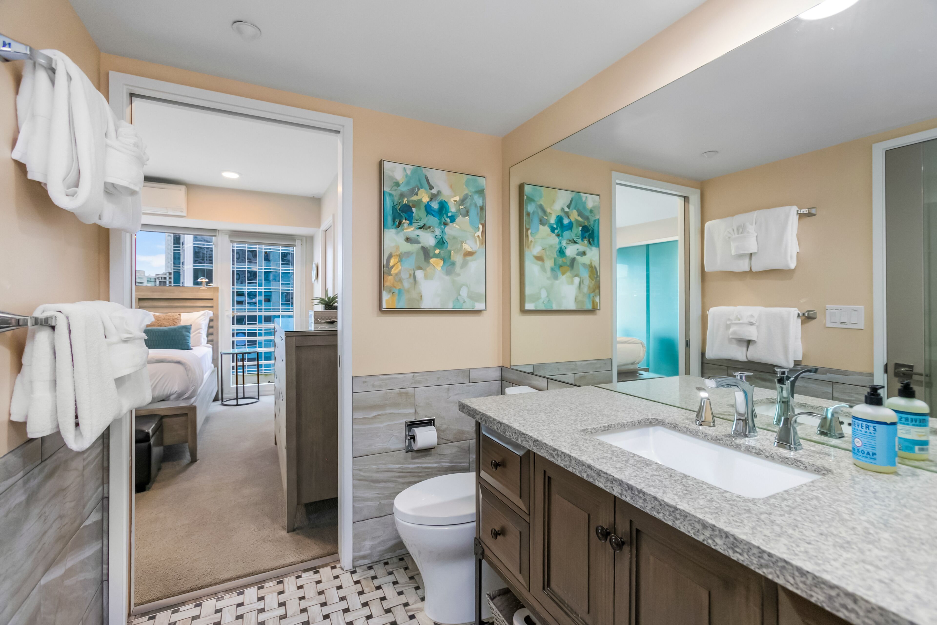 A modern bathroom with a large mirror, two sinks, and a toilet. There are towels hanging on a rack and a shower area visible through an open door.