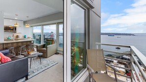 A modern apartment with a balcony overlooking a body of water, featuring a kitchen, seating area, and large windows. - NewMark Towers Water Wheel Sea Views (Seattle)