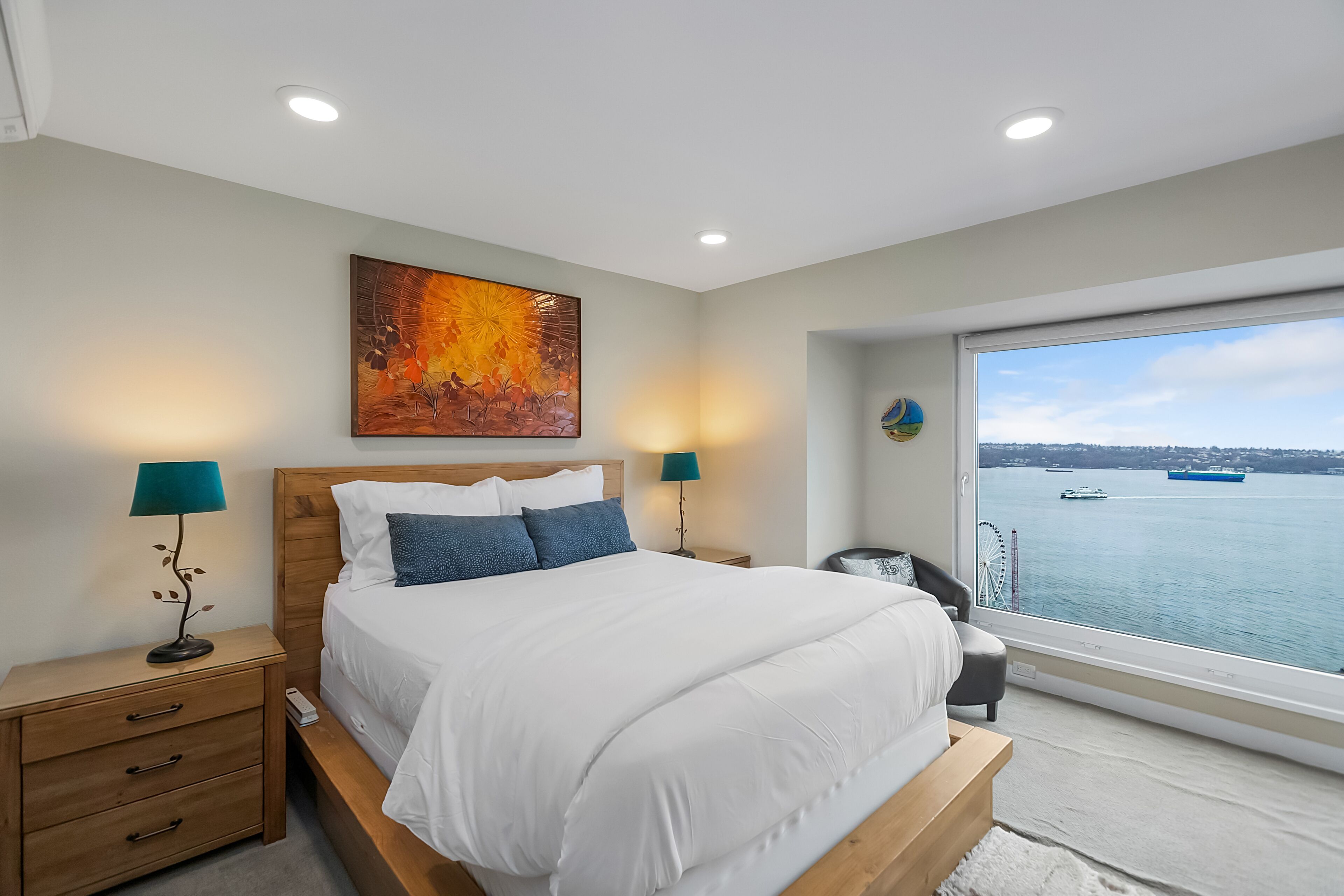 A bedroom with a large bed, two bedside tables with lamps, a painting on the wall, and a view of the sea through a large window.
