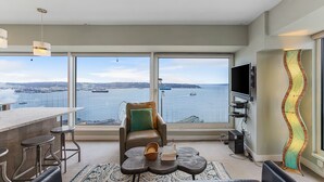 A modern living room with a large window offering a view of the water and ships, a leather sofa, a coffee table, and a TV mounted on the wall. - NewMark Towers Water Wheel Sea Views (Seattle)