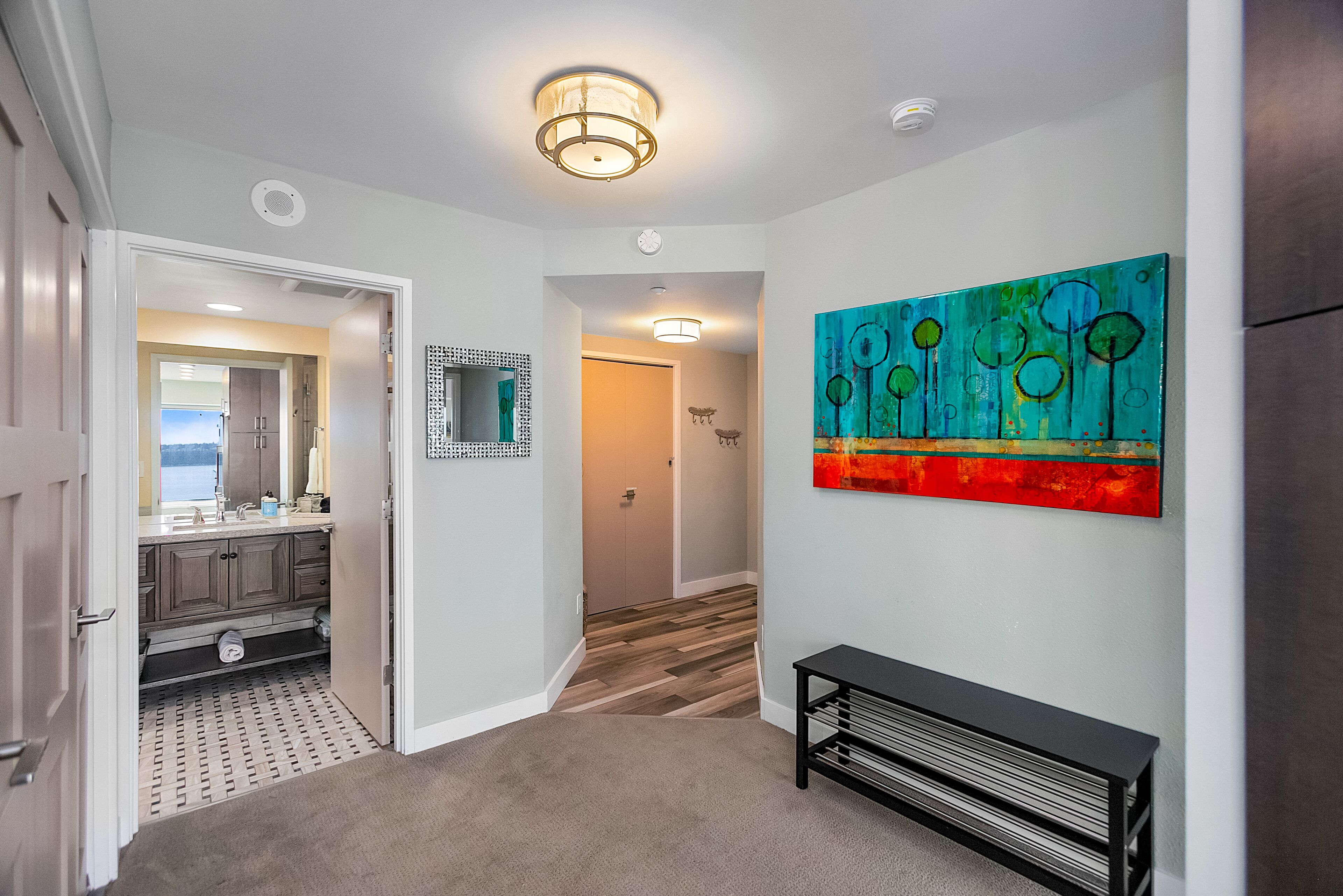 A hallway with a wooden floor, a bench, a wall-mounted artwork, and a door leading to a bathroom.