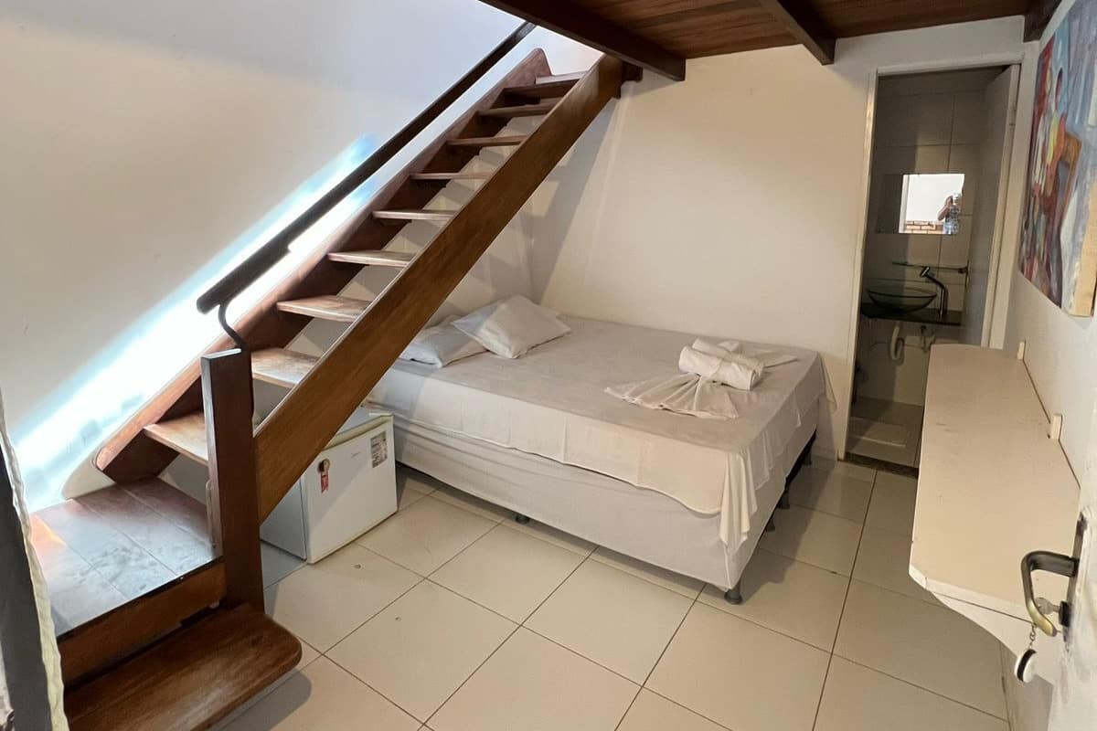 Ein kleiner, kompakter Raum mit einer Holztreppe, die zu einer oberen Ebene führt. Ein Einzelbett mit weißer Matratze und Kissen lehnt an der Wand. Der Raum hat einen Fliesenboden und eine weiße Decke.