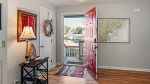 A hallway with a wooden floor, a red door, a map on the wall, a lamp, and a small table with a rug. - Tidal Wave Retreat (Beaufort)