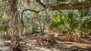 An outdoor dining area with a wooden table and chairs, surrounded by trees and a palm plant. - Tidal Wave Retreat (Beaufort)