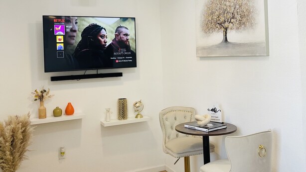 A modern living room with a flat-screen TV, a chandelier, a framed artwork, a white chair, and a small round table with a book and a sign.