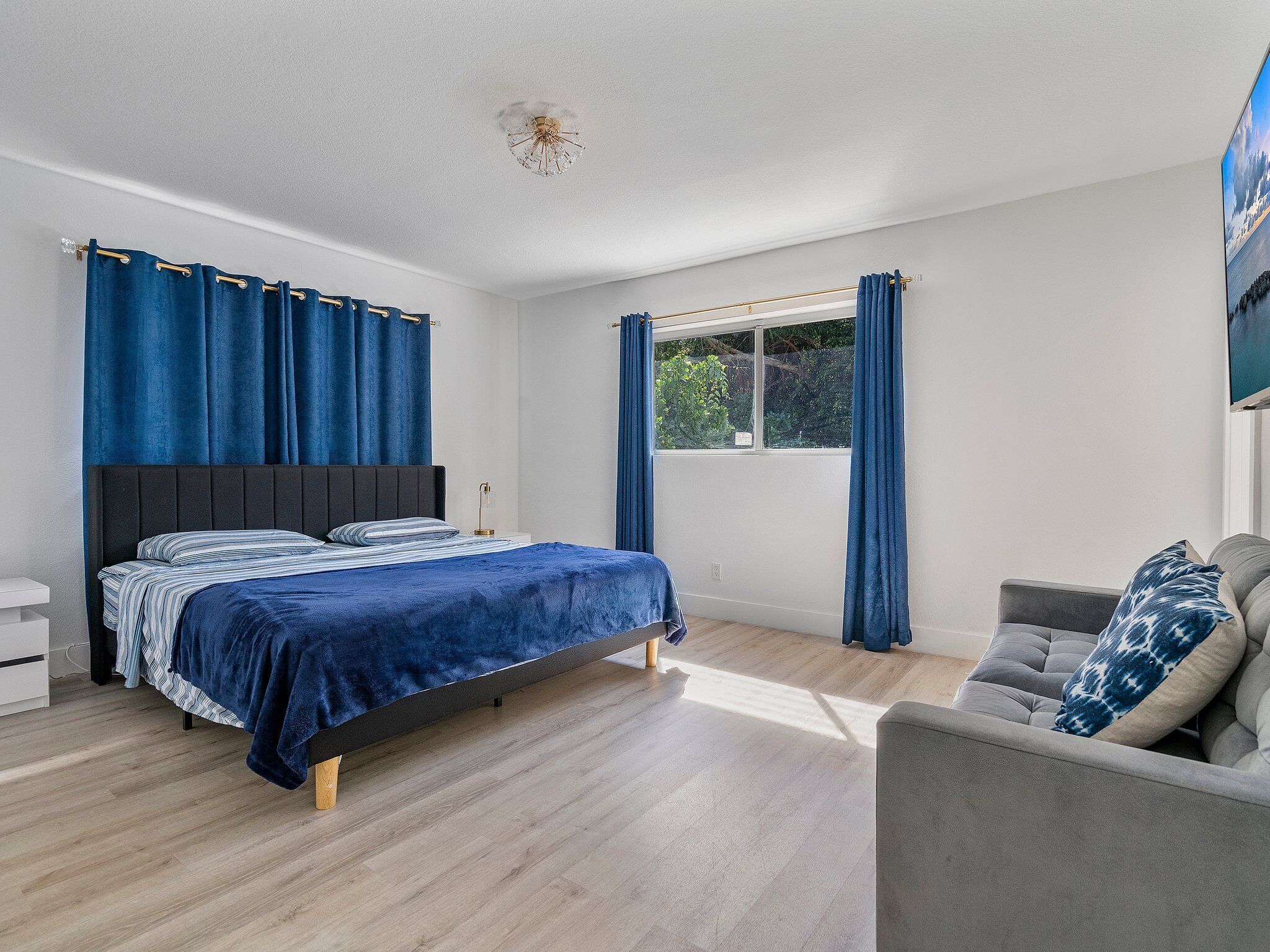 A modern bedroom with a bed, a nightstand, a grey sofa, and a window with curtains.