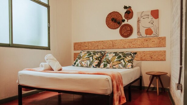 Een bed met een witte matras, een bloemenkussen en een deken. Aan de muur boven het bed hangen geweven manden.