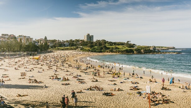 Una playa concurrida, gente tomando el sol y nadando, cielo despejado y edificios al fondo.