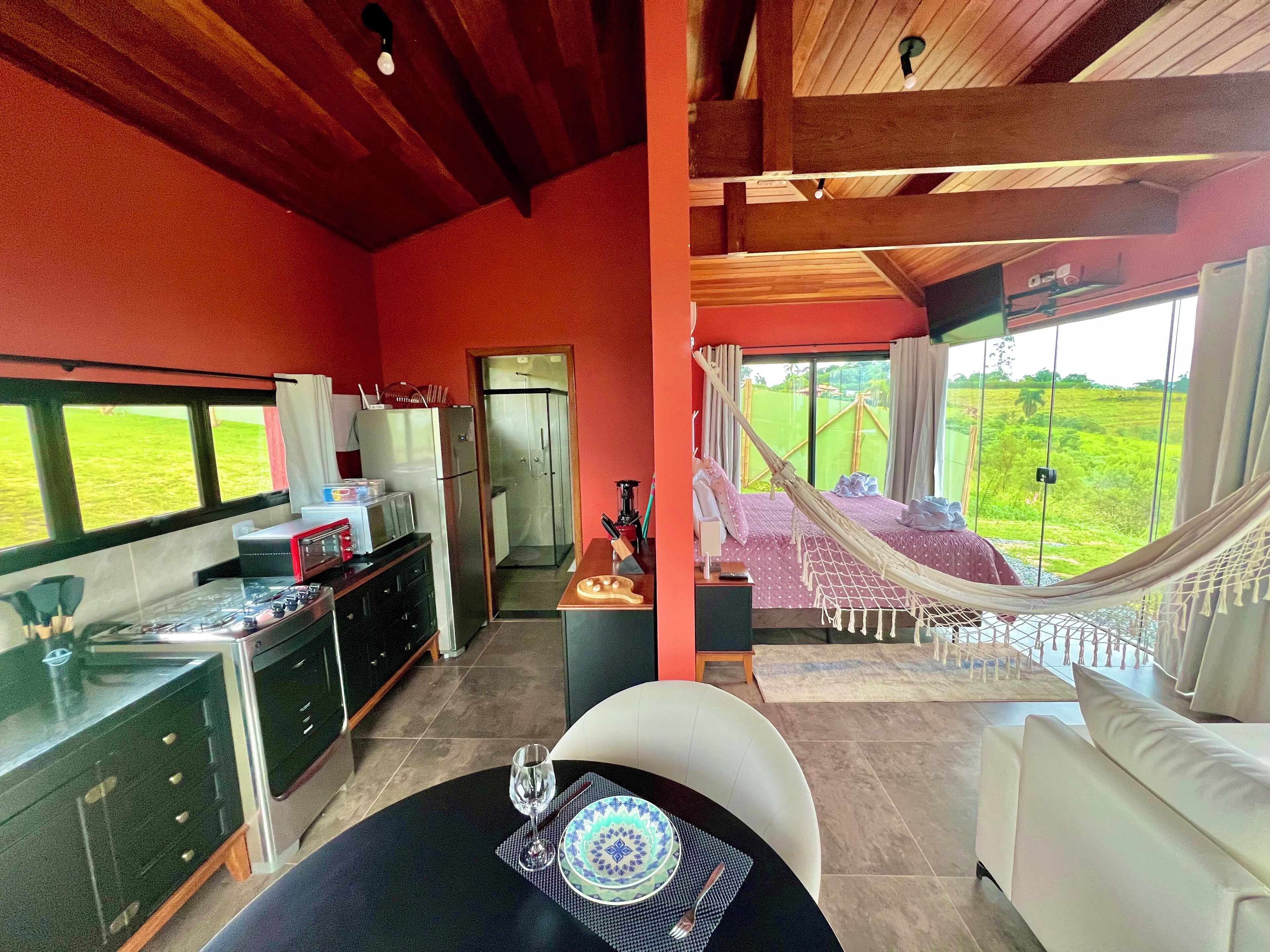 A modern kitchen with a gas stove, refrigerator, and microwave. There is a dining area with a table, chairs, and a glass of wine. A hammock is set up near a large window overlooking a green landscape.