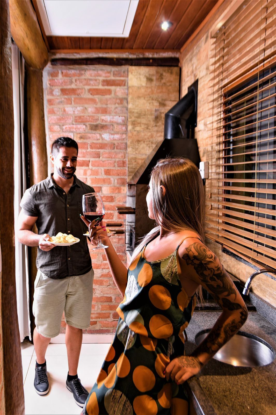 A man and a woman in a rustic kitchen with brick walls, a wooden ceiling, and a black stove.