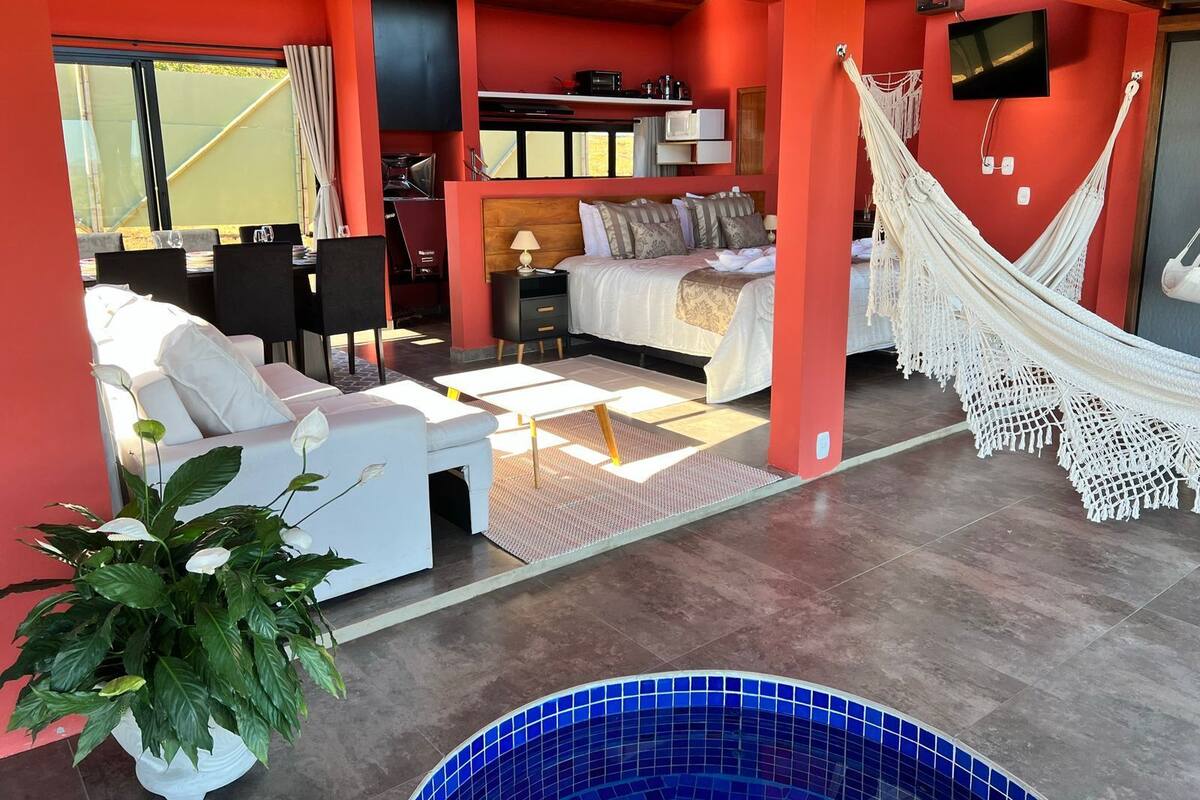 A modern hotel room with a bed, a sofa, a dining area, a TV, and a hammock.